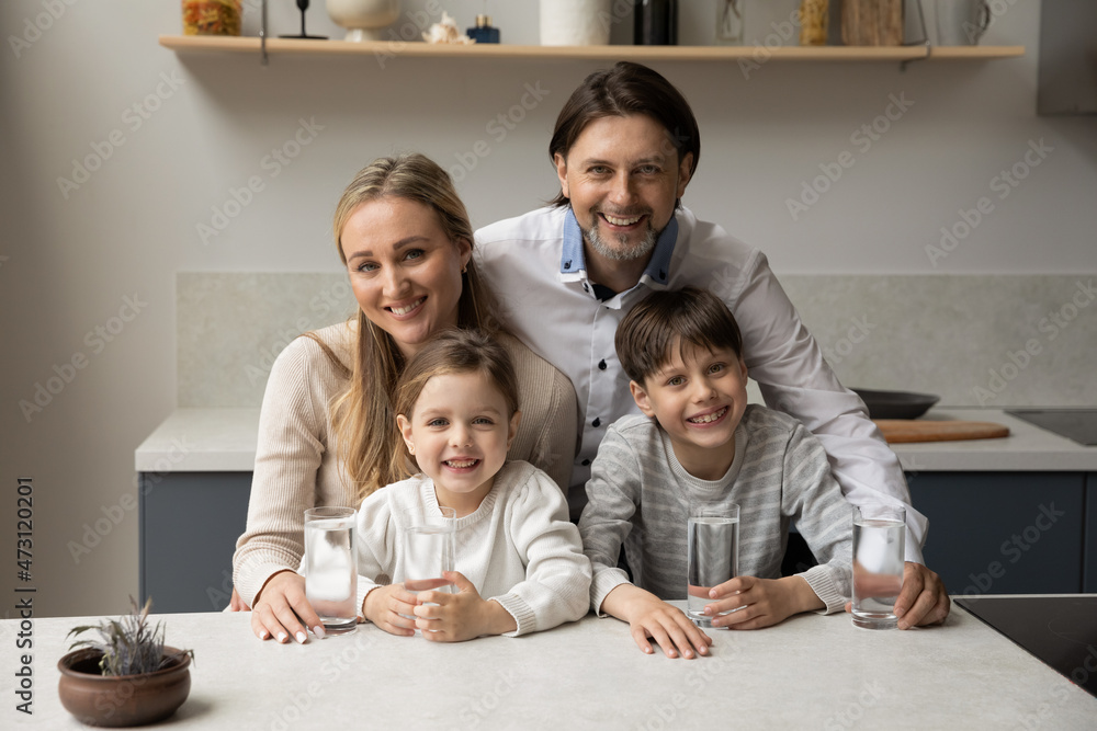 Portrait of smiling bonding young couple parents and little children siblings holding glasses of fresh pure mineral water in hands, refreshing in modern kitchen, daily healthcare family habit concept. Portrait of smiling bonding young couple parents and little children siblings holding glasses of fresh pure mineral water in hands, refreshing in modern kitchen, daily healthcare family habit concept.