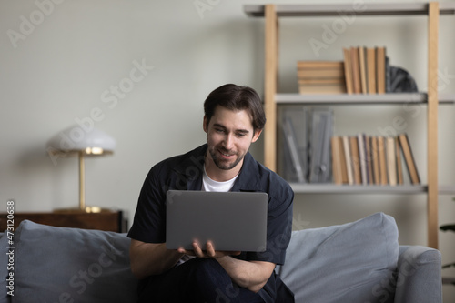 Happy smiling guy working at computer from home couch, holding portable laptop, typing, using internet app, online service on digital device for shopping, virtual learning, browsing