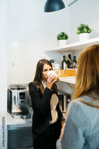 Two business woman drinking coffee, in a white mug, during a break at the coffee area in the office