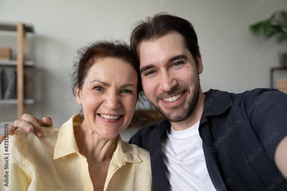 Happy adult son and mature mom hugging with love, affection, taking selfie, looking, smiling at camera, having fun, enjoying leisure at home together. Head shot portrait. Motherhood concept Happy adult son and mature mom hugging with love, affection, taking selfie, looking, smiling at camera, having fun, enjoying leisure at home together. Head shot portrait. Motherhood concept