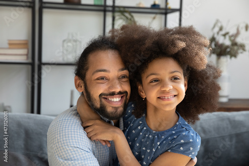 Head shot affectionate loving African American father cuddling little cute curly kid daughter, looking in distance, dreaming or visualizing future together at home, warm family relations concept.