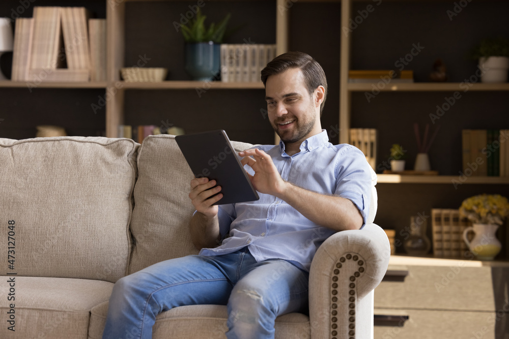 Happy young man holding digital device, resting on couch at home, using online virtual app on tablet, browsing internet, chatting, reading book, watching multimedia content, laughing