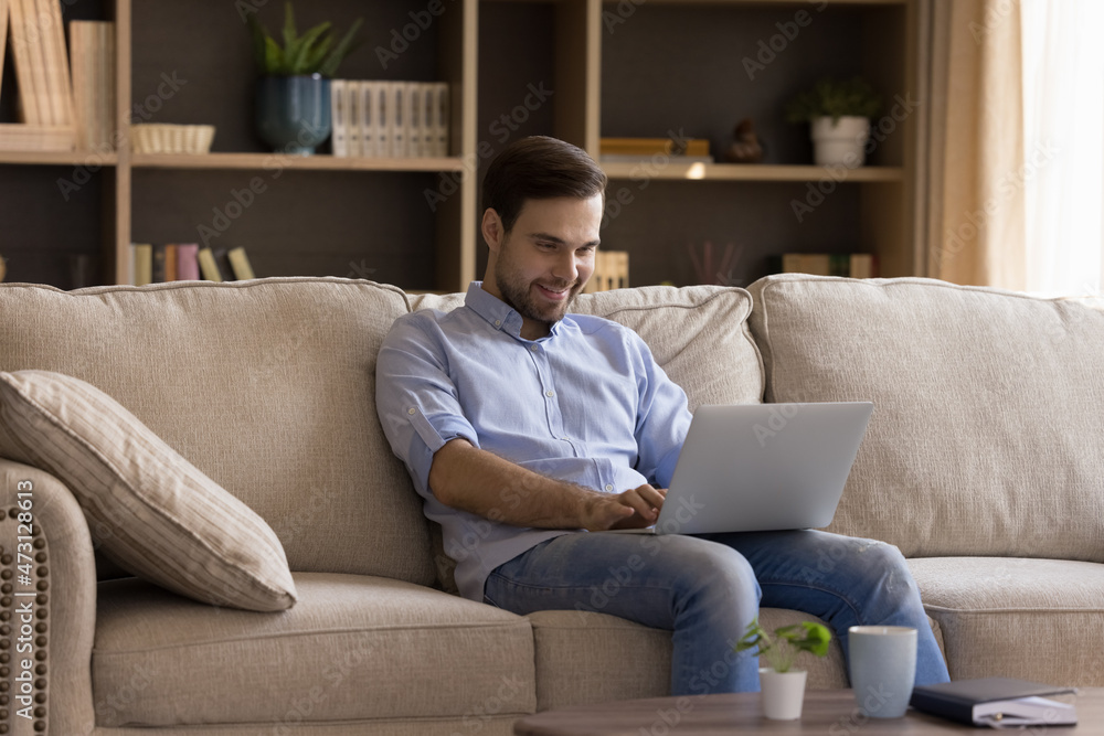 Happy millennial freelance worker using laptop at home, sitting on comfortable sofa, typing, smiling. Young business man working on project, chatting online, shopping, dating virtually
