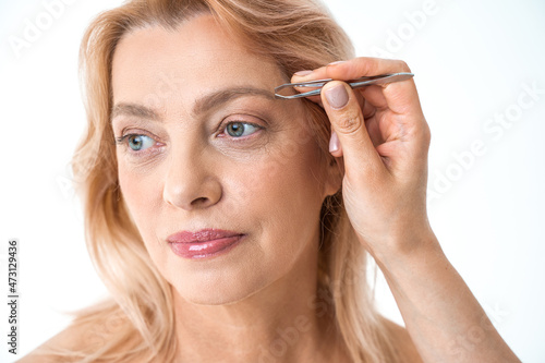 Woman correcting eyebrows with tweezers and looking away
