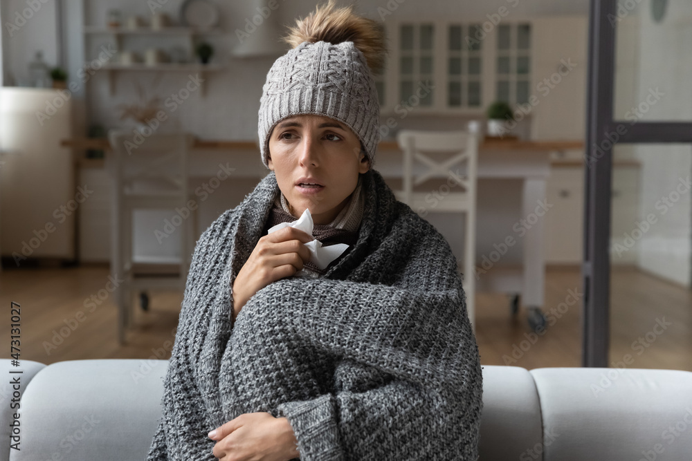 Sick woman covered with blanket caught cold, sitting on couch at home, unhealthy young female wearing cap feeling unwell, sneezing holding paper napkin, suffering from fever virus symptoms Sick woman covered with blanket caught cold, sitting on couch at home, unhealthy young female wearing cap feeling unwell, sneezing holding paper napkin, suffering from fever virus symptoms
