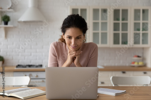 Smiling woman looking at laptop screen, working or studying online in modern kitchen at home, interested positive young female watching webinar or video training, reading good news in message