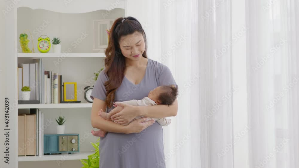 Beautiful Asian woman holding newborn baby in her arms standing in front of windows at cozy home.Happy infant baby sleep in mother arms safety and comfortable.Mom and Baby Concept Beautiful Asian woman holding newborn baby in her arms standing in front of windows at cozy home.Happy infant baby sleep in mother arms safety and comfortable.Mom and Baby Concept