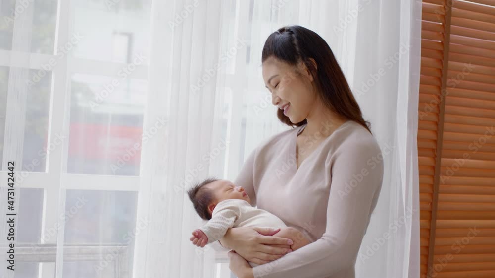 Beautiful Asian woman holding newborn baby in her arms standing in front of windows at cozy home.Happy infant baby sleep in mother arms safety and comfortable.Mom and Baby Concept Beautiful Asian woman holding newborn baby in her arms standing in front of windows at cozy home.Happy infant baby sleep in mother arms safety and comfortable.Mom and Baby Concept