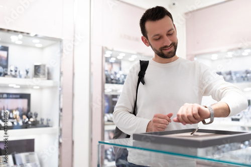 Photo of young positive happy handsome brunette man in a white sweatshirt chooses and tries on a stylish wrist watch in a store in a shopping center