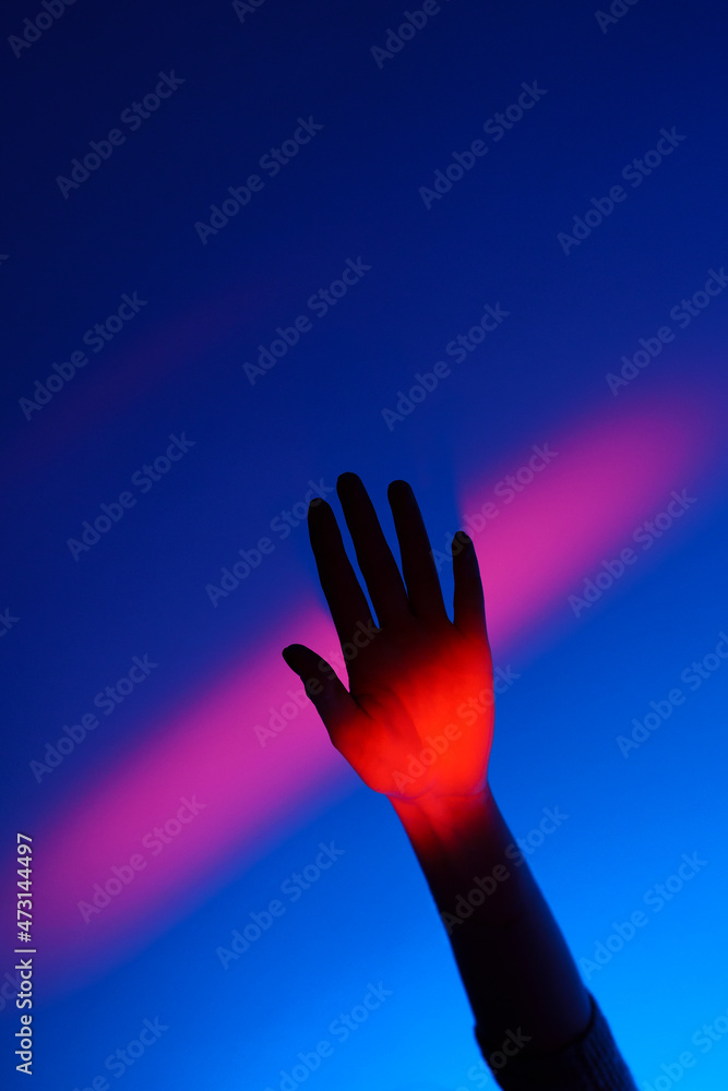 Closeup of anonymous woman arm gesturing with open hand over fashion blue wall, neon red stripe on wrist and background. Colorful light, minimalism concept. 
