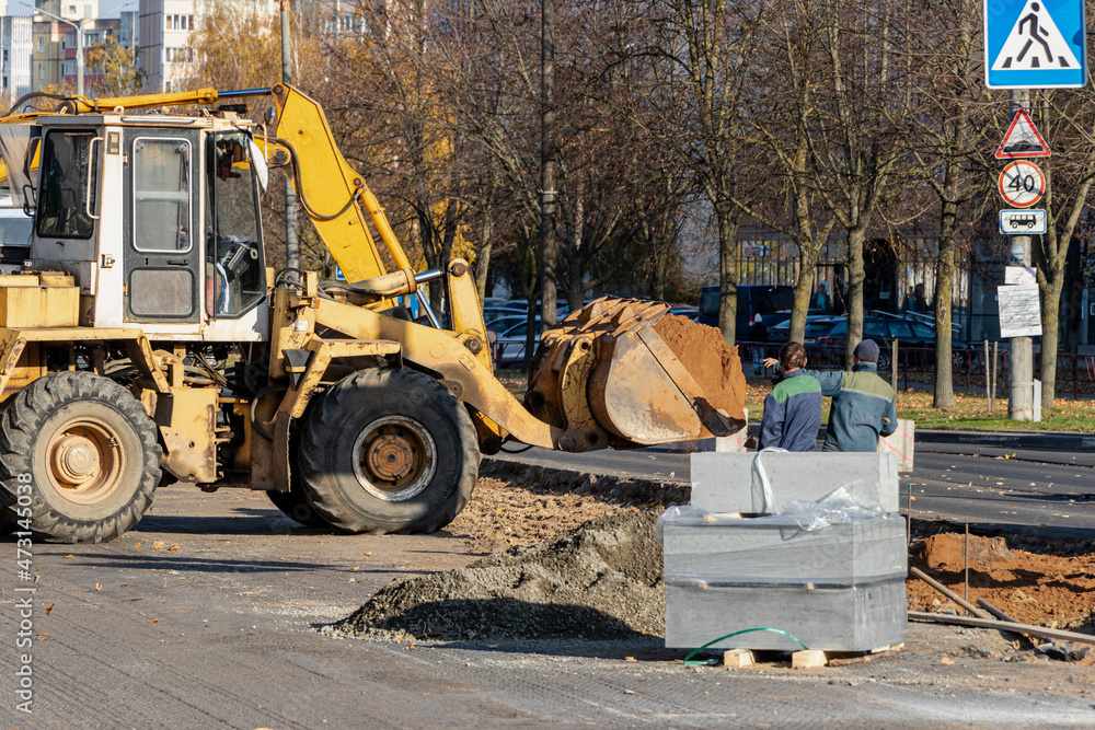 A heavy front loader transports earth in a bucket and grades the road. Road repair in the center of a modern city in autumn. Heavy construction machinery for moving soil.