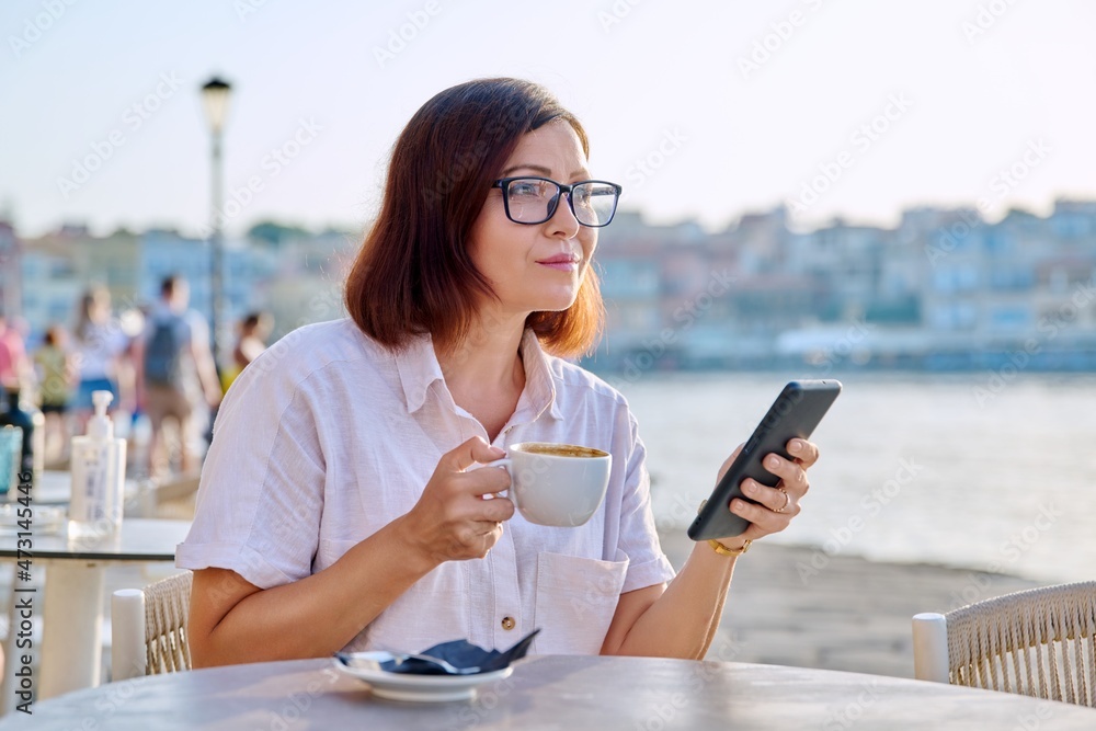 Mature woman in outdoor sidewalk cafe on seafront with cup of coffee using smartphone Mature woman in outdoor sidewalk cafe on seafront with cup of coffee using smartphone