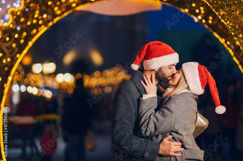 Christmas people kissing outdoors on New Year s eve. A young couple in love with Santa hats on heads standing on the street and kissing on the lips during the Christmas holidays.