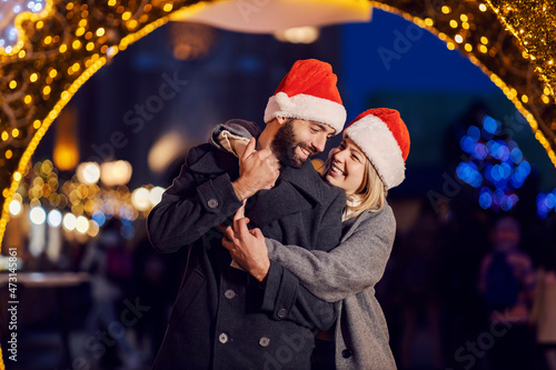 Romantic Christmas people hugging outdoors on New Year's eve. A happy couple in love with Santa hats on their heads is standing on the street on Christmas eve and hugging.