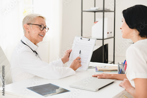 Smiling caucasian senior female doctor talking about health insurance test results diagnosis, giving prescriptions to female patient at hospital.