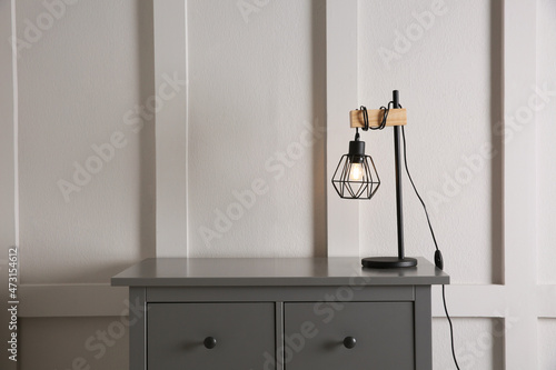 Wooden chest of drawers with modern lamp near white wall, space for text. Interior element