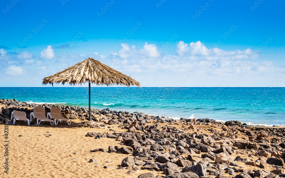 Sunny beach on Lanzarote island in Spain. Natural umbrella from dry palm leaves and sun chairs on sandy beach with lava rocks, turquoise ocean waves on Canary Islands