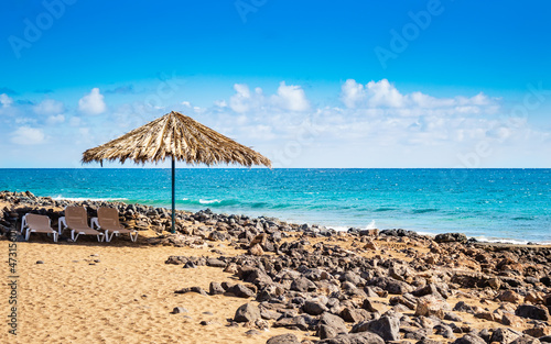 Sunny beach on Lanzarote island in Spain. Natural umbrella from dry palm leaves and sun chairs on sandy beach with lava rocks, turquoise ocean waves on Canary Islands © Julia Lavrinenko