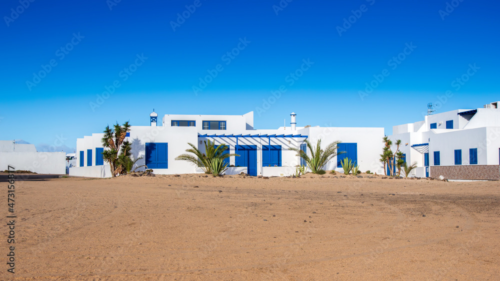 Typical canarian white and blue houses in Caleta del Sebo town on la Graciosa island, near Lanzarote in Spain. Beautiful traditional home on deserted sandy terrain near sea under bright blue sky Typical canarian white and blue houses in Caleta del Sebo town on la Graciosa island, near Lanzarote in Spain. Beautiful traditional home on deserted sandy terrain near sea under bright blue sky