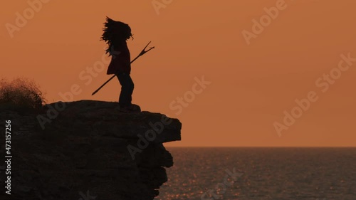 Silhouette of Native American approaches edge of cliff of sea coast and looks into distance on background of sunset sky. photo