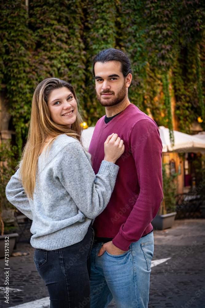 Fototapeta premium Young couple traveling to Rome. Beautiful blonde woman hugs her boyfriend. In the background, a street in the historic center with an ivy-covered building.