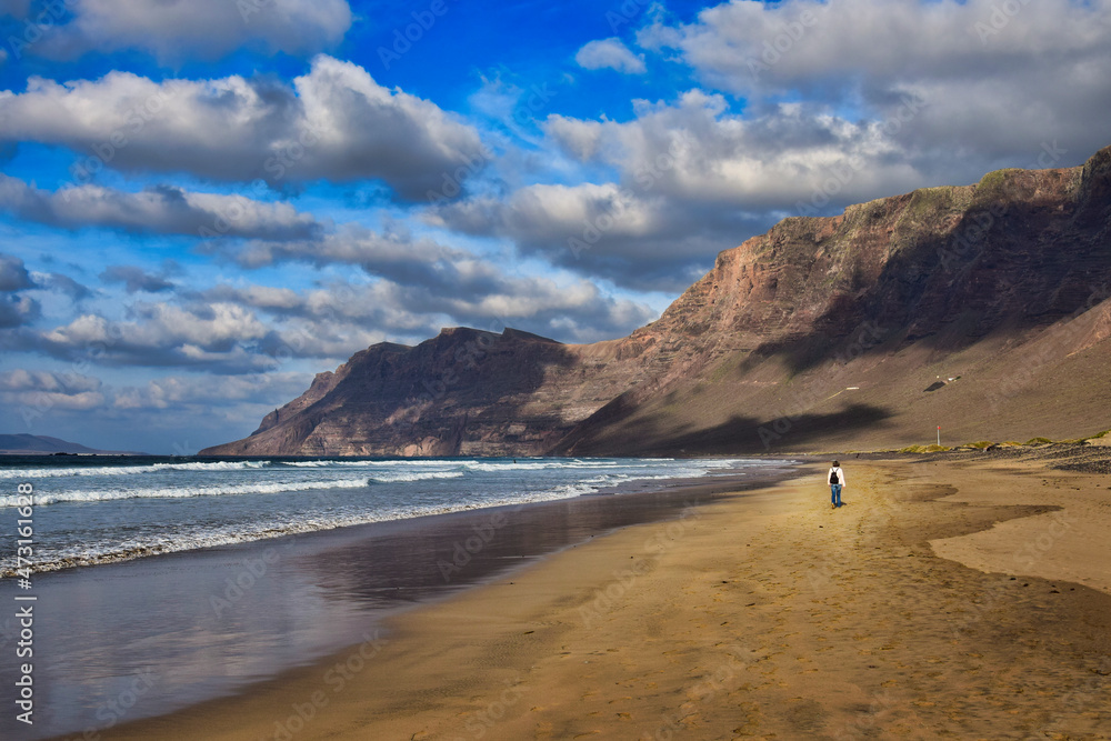 Beautiful Famara Beach, Lanzarote, Spain. Mountains in the background. A woman walking along the sandy beach. A blue sky with some clouds.
