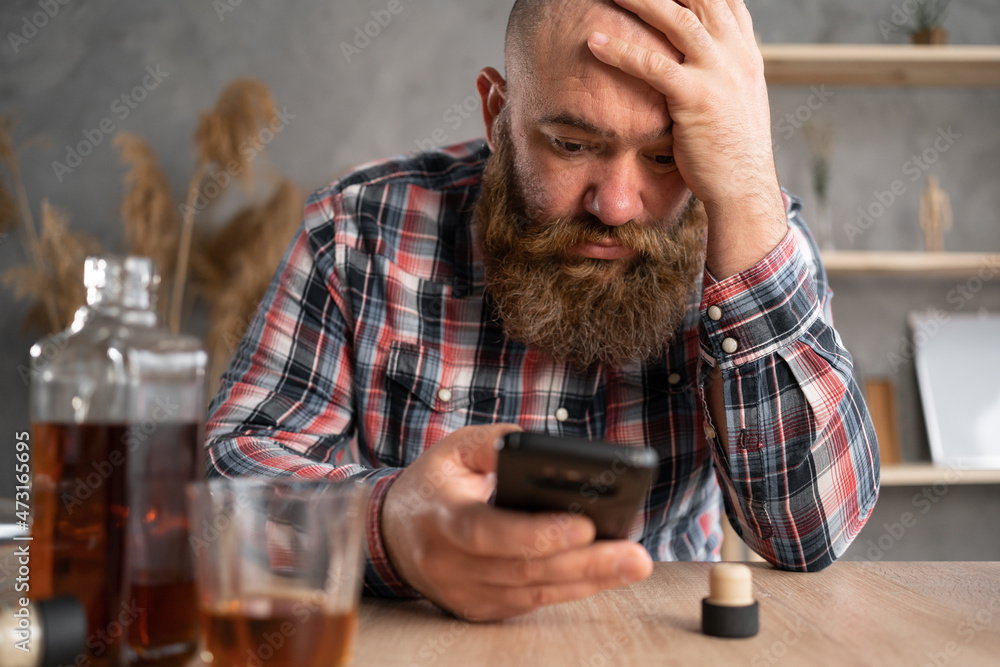 alcoholism, alcohol addiction and people concept a bearded alcoholic man with a smartphone in casual clothes sits at a table near a bottle of whiskey and a glass.