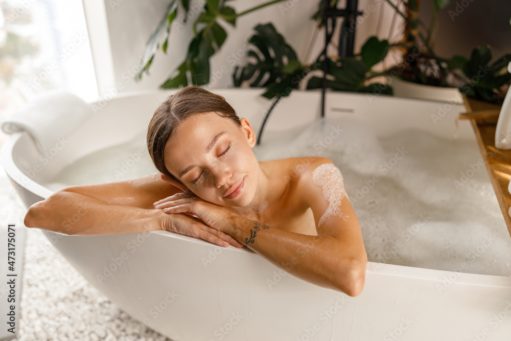 Obraz premium Attractive young woman feeling tired, leaning on bathtub side, relaxing with eyes closed at spa resort. Wellness, beauty and care concept