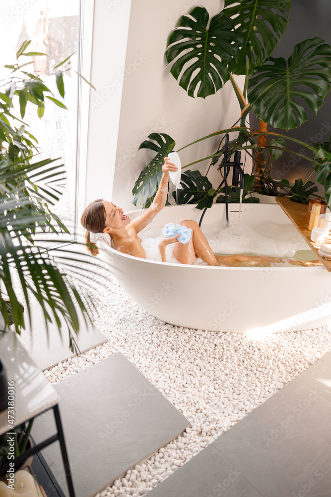 Cheerful young woman having fun while pouring shower gel on loofah sponge, taking bath in modern bathroom decorated with tropical plants. Spa, wellness, body care concept