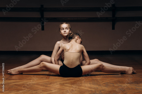 Image of two beautiful young ballerinas doing the splits, stretching their legs in a pose on stage, in the studio. Elegant performer, flexibility of concept, strength discipline, physical work.
