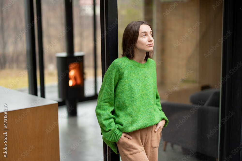 Young woman stands at window of a modern house on nature and look away. Concept of comfort and happy modern life. Woman wearing green sweater Young woman stands at window of a modern house on nature and look away. Concept of comfort and happy modern life. Woman wearing green sweater