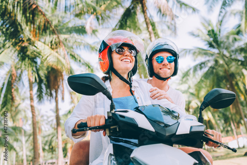 Happy smiling couple travelers riding motorbike scooter in safety helmets during tropical vacation under palm trees on Ko Samui island , Thailand