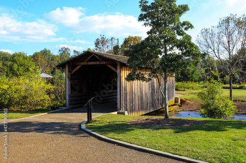 a shot of a wooden covered bridge over a river surrounded by lush green and autumn colored trees with lush green grass, blue sky and clouds at sunset at Garrard Landing Park in Alpharetta Georgia USA	 photo