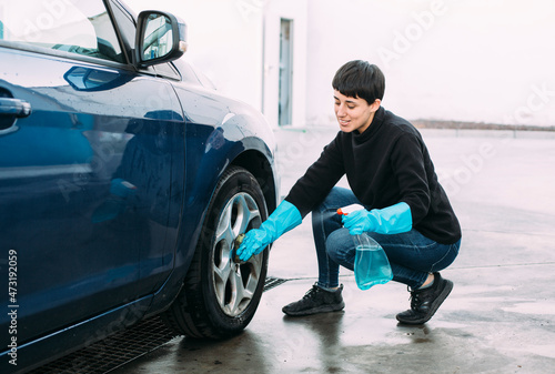 Young woman cleaning the tire of a blue car using a sponge and a spray while wearing blue rubber gloves. Concept of self-service car washing. 