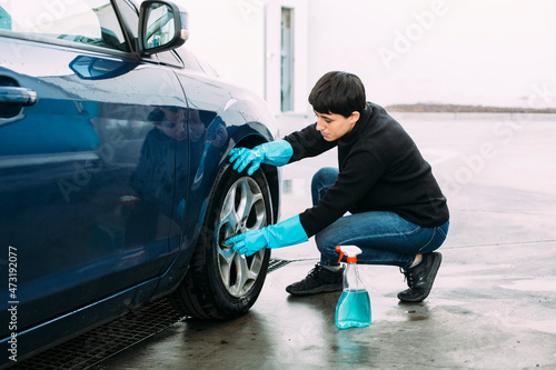 Young woman cleaning the tire of a blue car using a sponge and a spray while wearing blue rubber gloves. Concept of self-service car washing. 