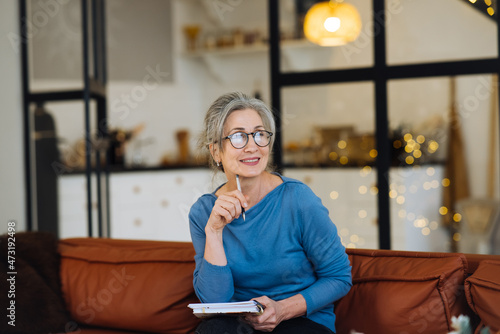 Happy senior woman in glasses thinking and writes in a diary home
