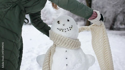 Woman dresses snowman in Christmas Santa hat, scarf and attaching fir branches in hands. Outdoors activities in snowy winter park