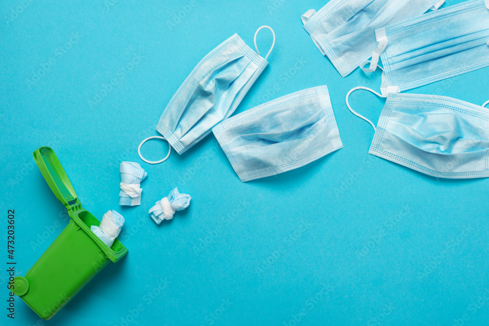 Throwing away a medical mask in a green container for recycling on a blue background. Blue face masks after use. The use of medical masks Throwing away a medical mask in a green container for recycling on a blue background. Blue face masks after use. The use of medical masks