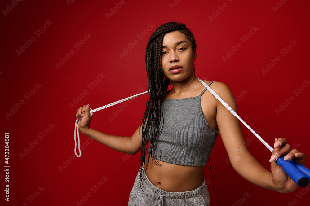African woman with stylish dreadlocks and fit body posing against red background with jumping rope. Cardio workout and fitness training concept with copy space for advertisement African woman with stylish dreadlocks and fit body posing against red background with jumping rope. Cardio workout and fitness training concept with copy space for advertisement