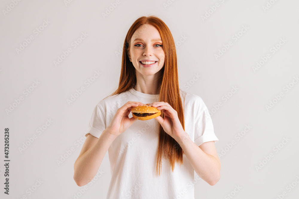 Studio portrait of cheerful attractive redhead young woman holding tasty burger and looking at camera standing on white isolated background. Happy cute female eating delicious hamburger.