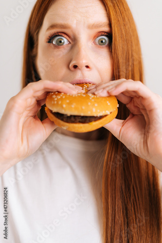 Close-up vertical shot of surprised cute young woman with eyes wide open pleasing bite of appetizing delicious burger looking at camera  standing on white isolated background in studio  front view.