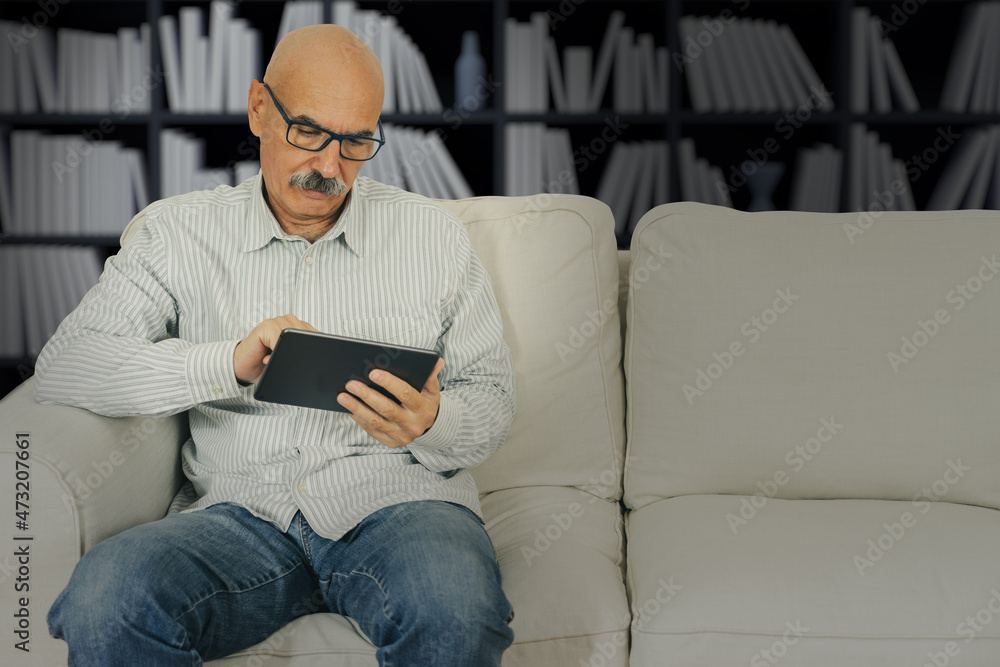 Senior Male Reading in a Digital Tablet Sitting at the Sofa at the House Living Room Senior Male Reading in a Digital Tablet Sitting at the Sofa at the House Living Room