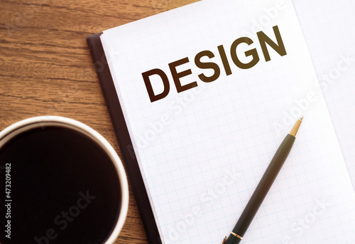 The word Design in a notebook on the office table next to glasses, a cup of coffee and a pencil
