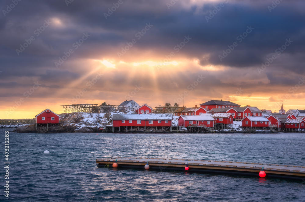 Naklejka premium Norwegian fishing village and sea coast at sunset. Lofoten islands, Norway. Winter landscape with red houses, blue water, dramatic cloudy sky with golden sun rays. Norwegian traditional red rorbu