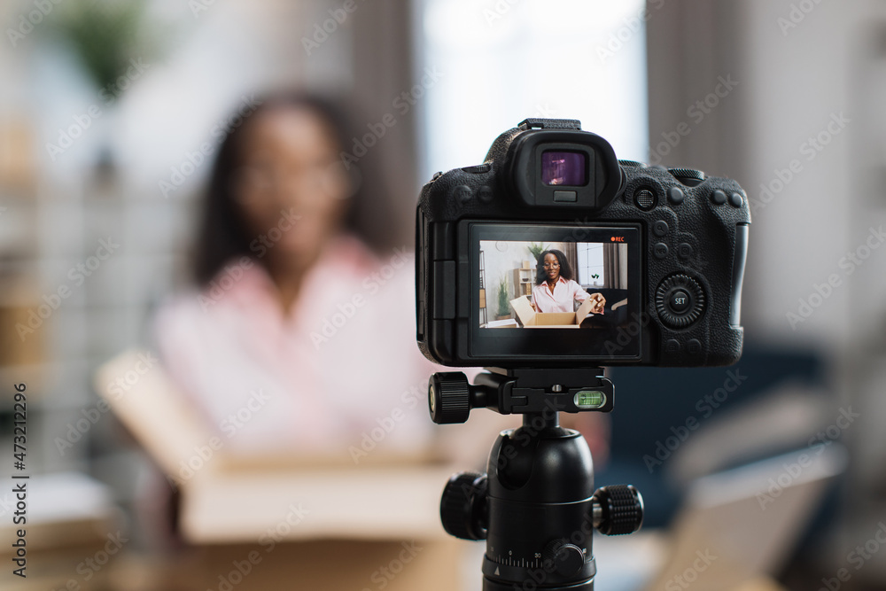 African american woman unboxing parcel with ordered goods while sitting at home and recording video blog. Female influencer using modern digital camera for creating new content. African american woman unboxing parcel with ordered goods while sitting at home and recording video blog. Female influencer using modern digital camera for creating new content.