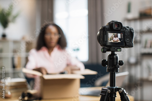 Blur background of young african woman unboxing parcel and recording video blog. Focus on modern digital camera. Concept of social networks technology.