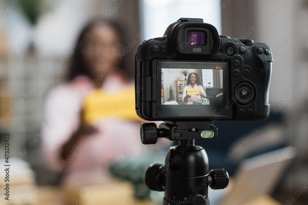 Close up of modern video camera with african american woman on screen. Female blogger unboxing holidays presents while sitting at desk. Blur background.