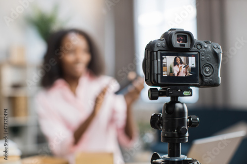 Blur background of young african woman recording video blog while unboxing new modern smartphone. Focus on screen of digital camera fixed on tripod.