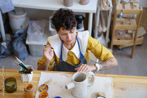 Craft artist at work. Focused woman artisian in pottery studio, drawing sketch on clay handmade teapot before applying paint and making ornament. Female ceramist producing earthenware kitchenware photo