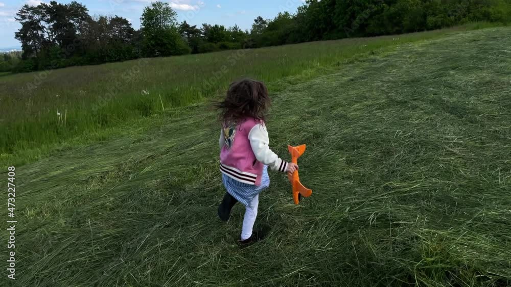 A young little girl running on a grassland with a toy plane in her hand. girl is running out of joy and suddenly falls down.